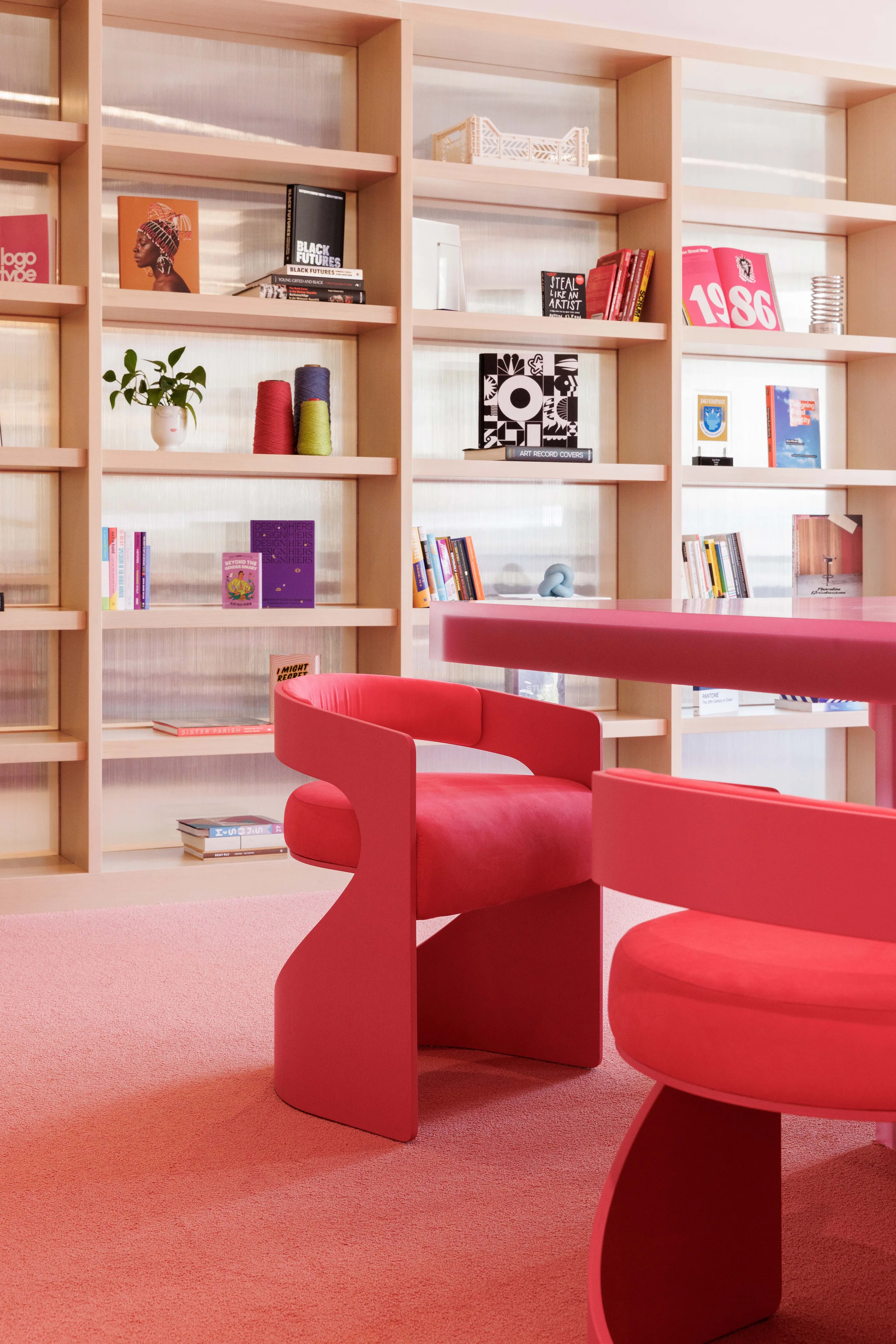 Red sculptural chairs on a pink carpet in front of tall shelves with books and objects.