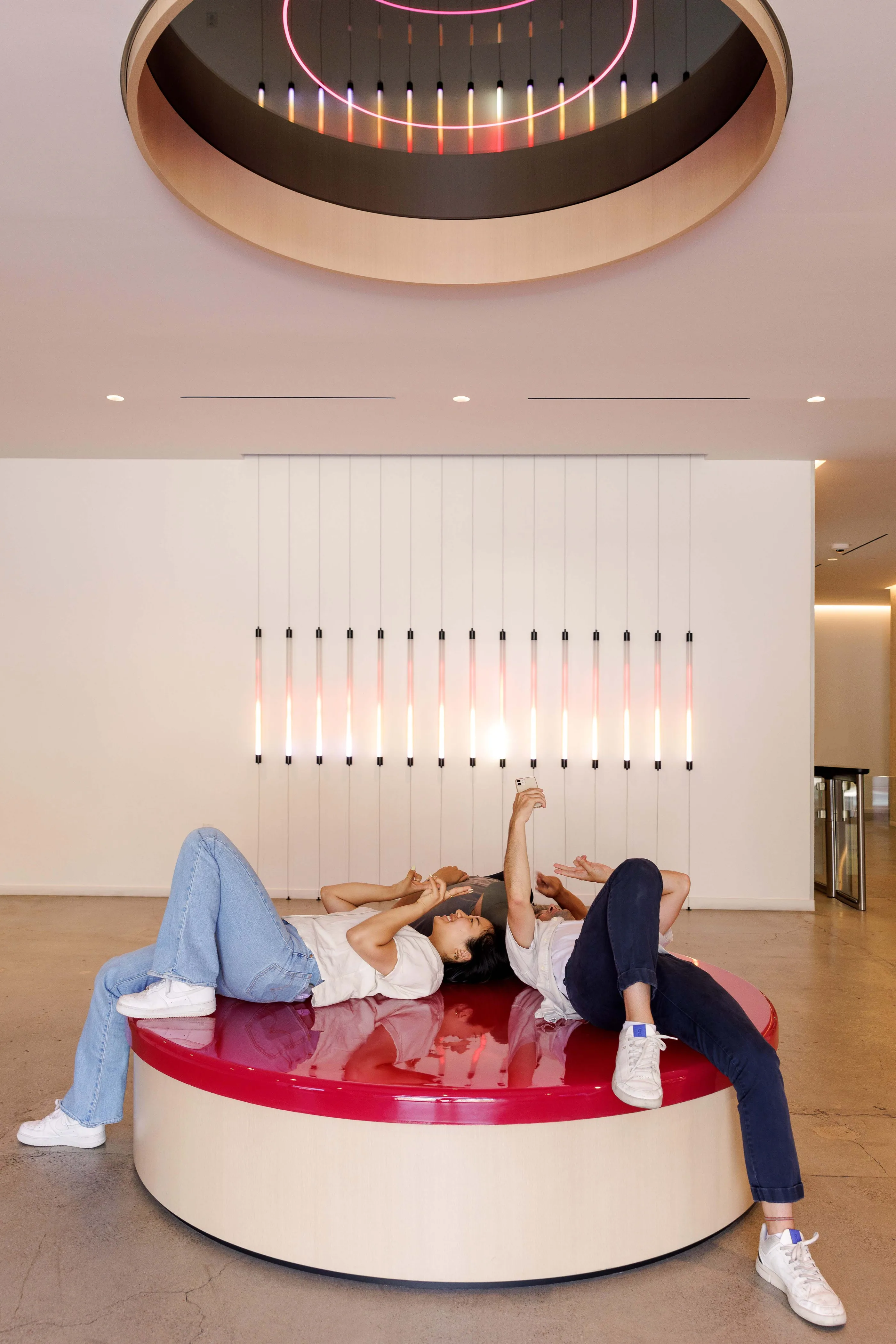 Lobby with a round red upholstered seat, a circular light fixture above, and vertical pink light tubes on the wall behind.